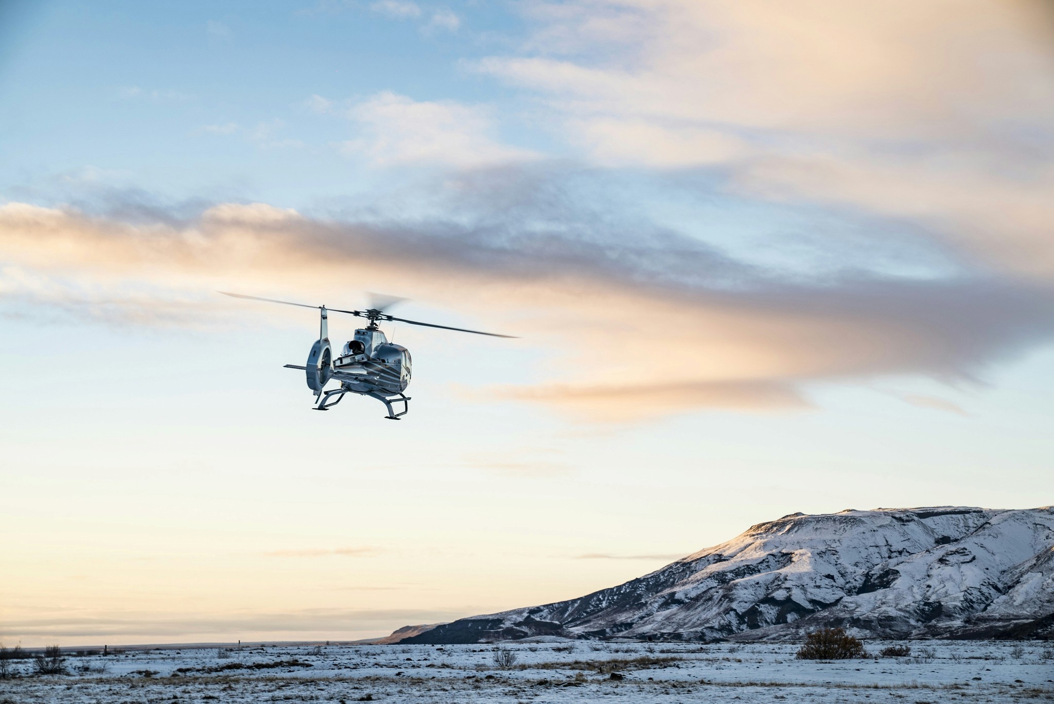 Helicopter flying over a snowy Icelandic landscape beneath a pinkish sky.